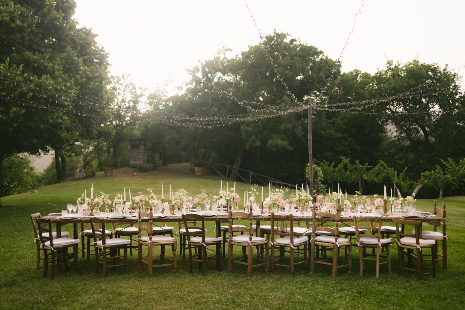 A long table with candles and chairs set on a lawn under strings of lights and trees