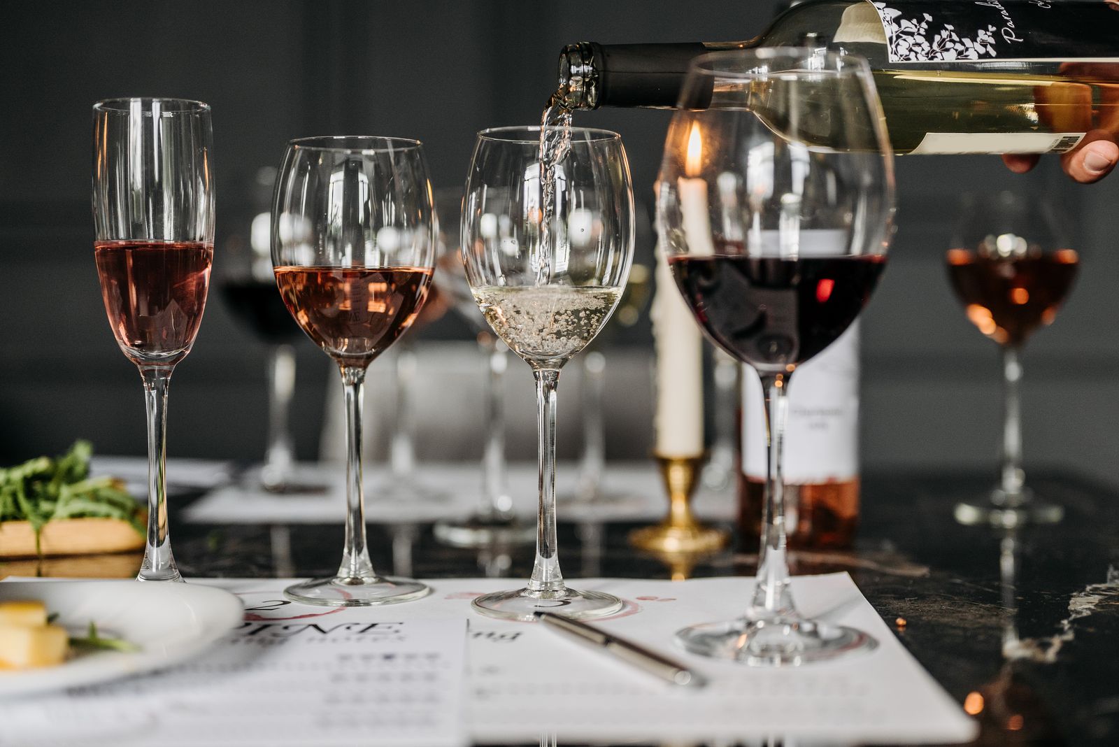 White wine being poured into a glass beside glasses of red and rosé on a tasting table set with tasting notes